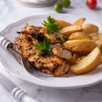 Baked chicken tenders on a white plate with potato wedges, a garnish of fresh herbs, and a fork on the plate.