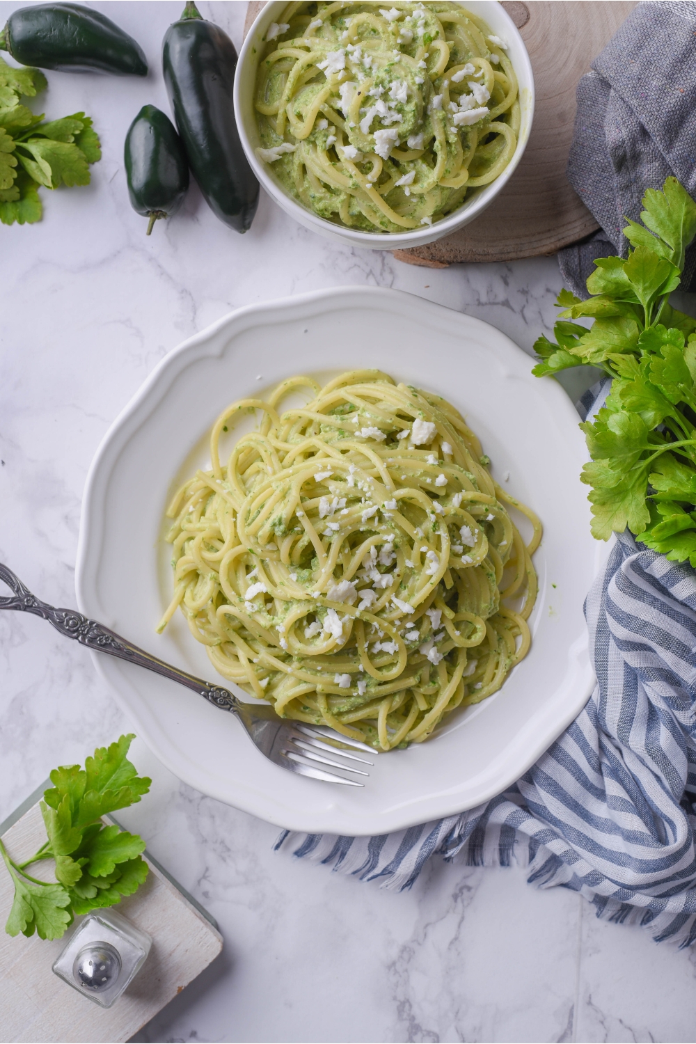 Overhead view of a bed of green spaghetti on a white plate topped with Cotija. There's a fork on the plate and a second bowl of pasta next to the plate.