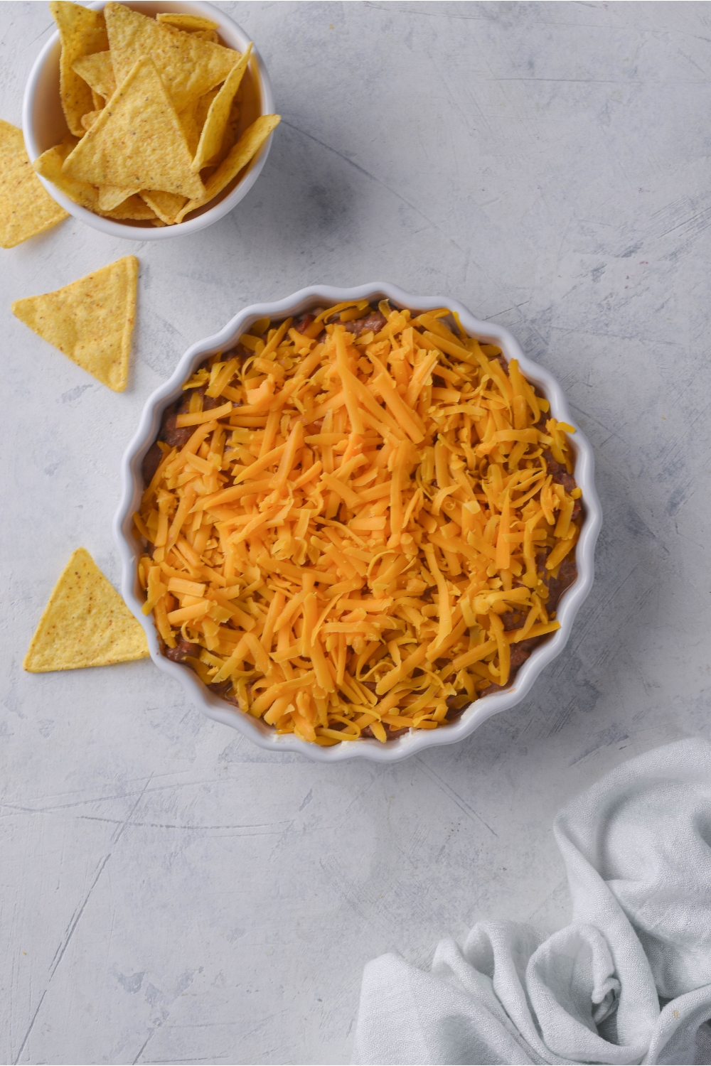 White serving dish with skyline chili dip and a layer of unmelted shredded cheese on top. Next to the chili dip is a smaller bowl of tortilla chips with some chips spilling over.