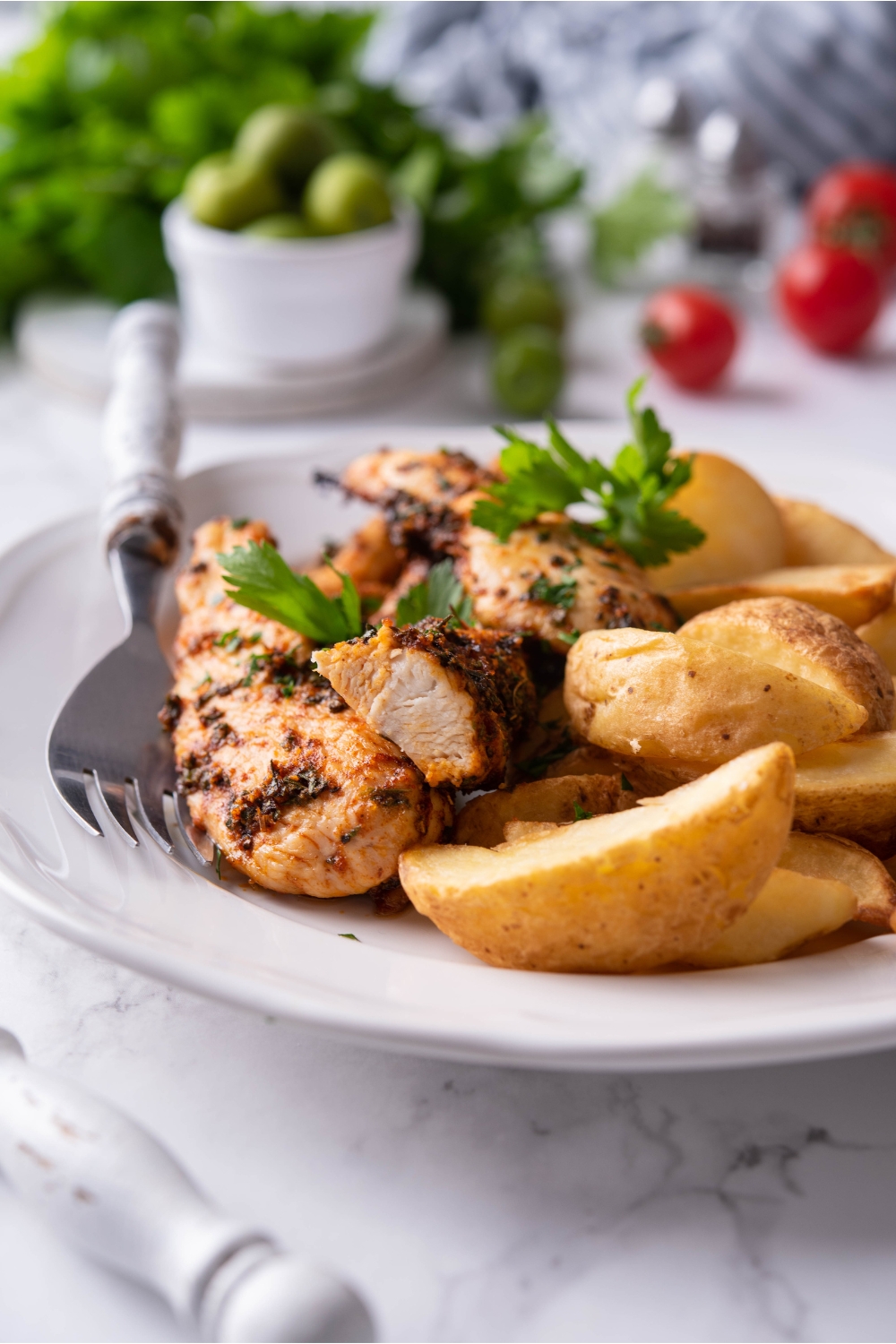 Baked chicken tenders on a white plate with potato wedges, a garnish of fresh herbs, and a fork on the plate.