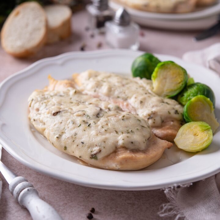 Baked chicken breasts covered in cream of chicken soup, on a white plate with a side of brussels sprouts and a set of silverware next to the chicken.