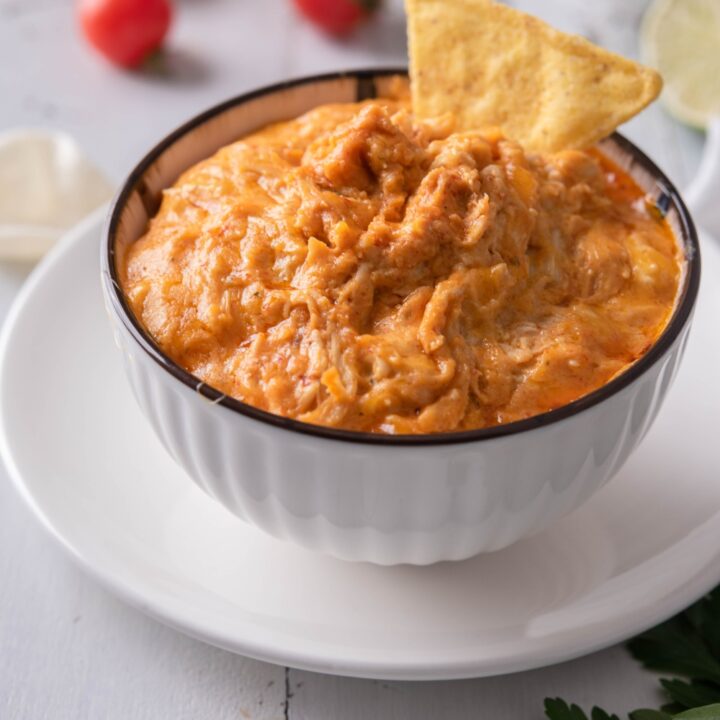 Buffalo chicken dip in a white serving bowl atop a white plate. There is a tortilla chip sticking out of the dip and in the background are cherry tomatoes and potato chips.