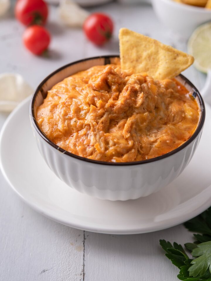 Buffalo chicken dip in a white serving bowl atop a white plate. There is a tortilla chip sticking out of the dip and in the background are cherry tomatoes and potato chips.