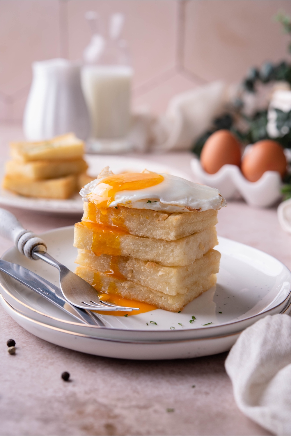 Four grit cakes stacked evenly on top of each other with a sunnyside up egg and the yolk oozing over the grit cakes. There is a set of silverware on the plate.