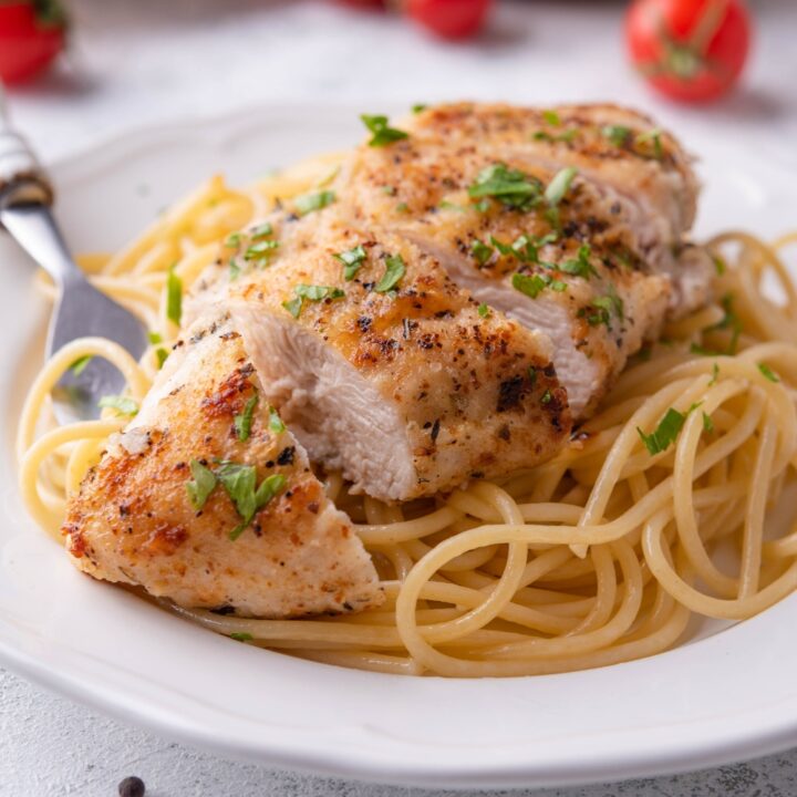A sliced pan-fried chicken breast atop a bed of spaghetti noodles on a white plate with a fork on the plate. The chicken is garnished with fresh green herbs.