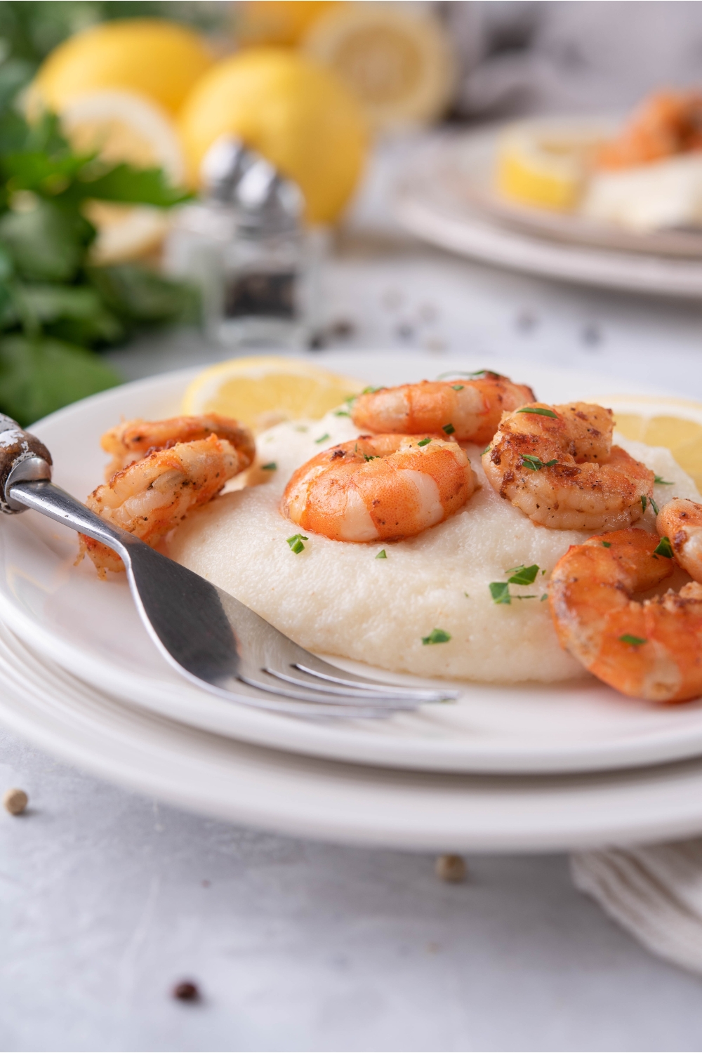 Pan seared shrimp atop grits with a garnish of fresh herbs and a lemon wedge. The plate of shrimp is on a second plate and there is a fork on the plate. In the background are more lemons and a second serving of shrimp.