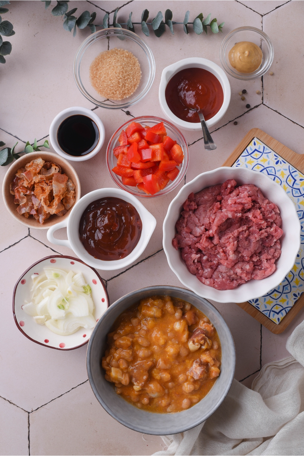 An assortment of ingredients including bowls of ground beef, diced peppers, barbecue sauce, diced onion, and canned beans.