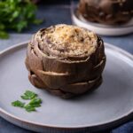 One stuffed artichoke topped with bread crumbs on a blue plate with a sprig of parsley on the plate.