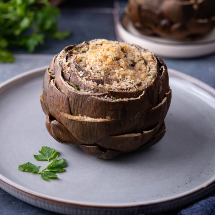 One stuffed artichoke topped with bread crumbs on a blue plate with a sprig of parsley on the plate.