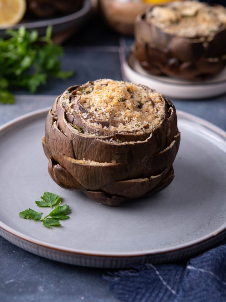 One stuffed artichoke topped with bread crumbs on a blue plate with a sprig of parsley on the plate.