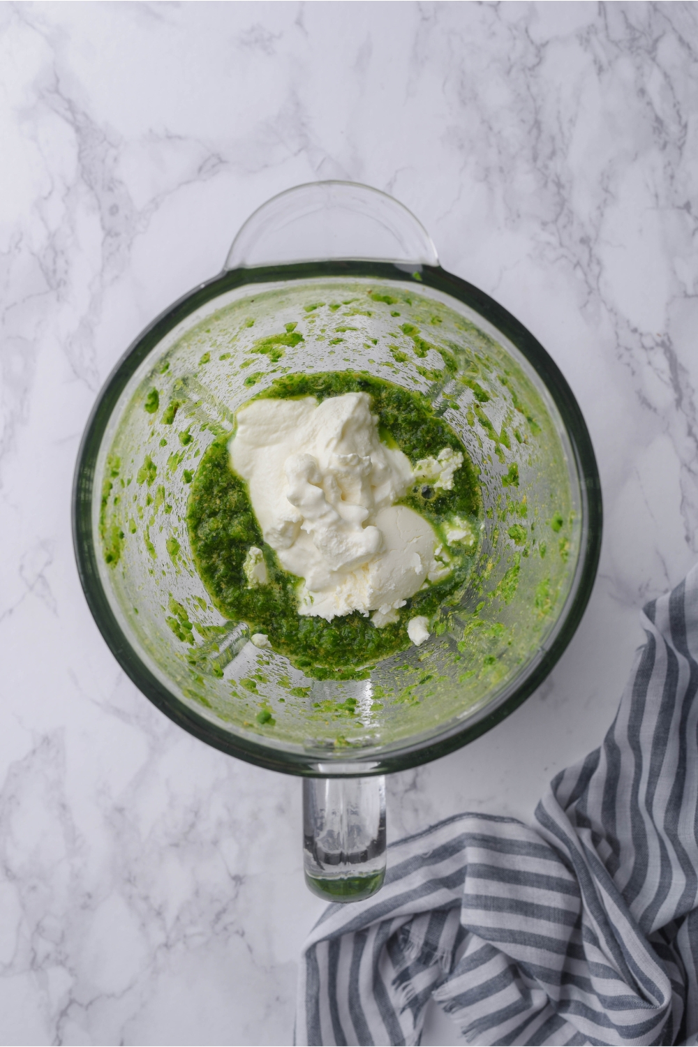 A food processor filled with a blended green sauce and a dollop of cream cheese that has not yet been mixed together with the green sauce.