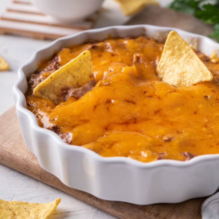 Skyline chili dip in a white serving dish with two tortilla chips sticking out of the dip. The serving dish is on a wood board and there is a bowl of tortilla chips in the background.
