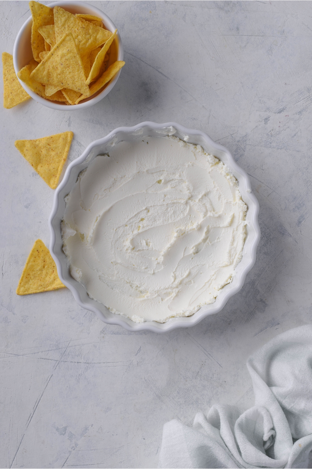 White serving dish with a smooth layer of cream cheese. Next to the bowl of chili dip is a smaller bowl of tortilla chips with some chips falling out onto the counter.