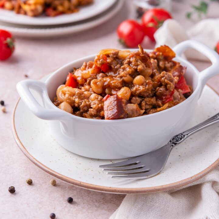Southern baked beans with ground beef and bacon in a white baking dish atop a white plate. There is a fork on the plate and in the background is a serving of baked beans with ground beef on a plate.