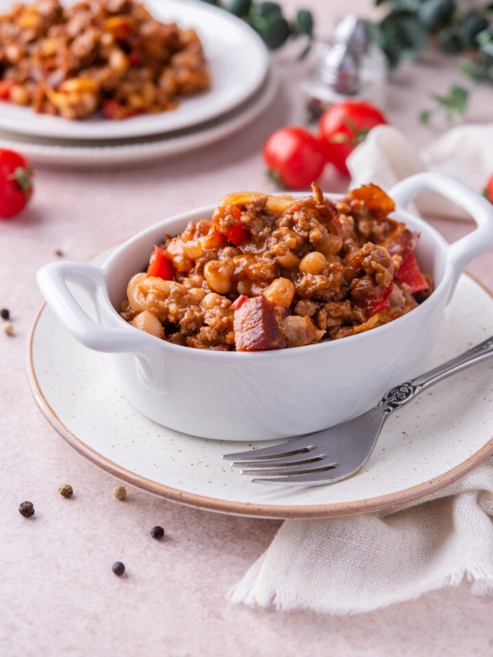 Southern baked beans with ground beef and bacon in a white baking dish atop a white plate. There is a fork on the plate and in the background is a serving of baked beans with ground beef on a plate.