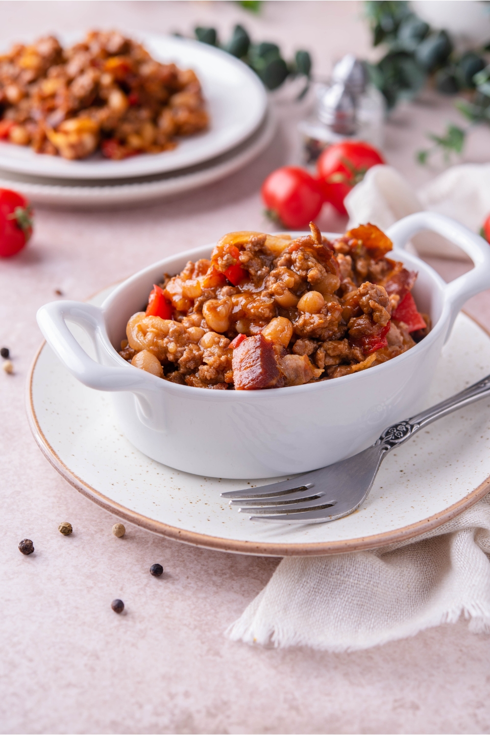 Southern baked beans with ground beef and bacon in a white baking dish atop a white plate. There is a fork on the plate and in the background is a serving of baked beans with ground beef on a plate.