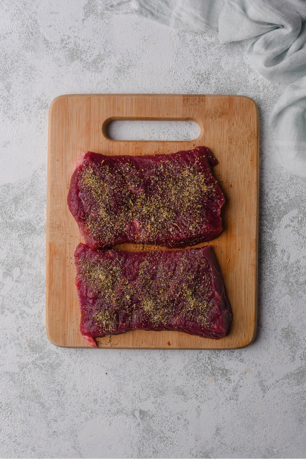 Two raw seasoned flat iron steaks on a wood cutting board.