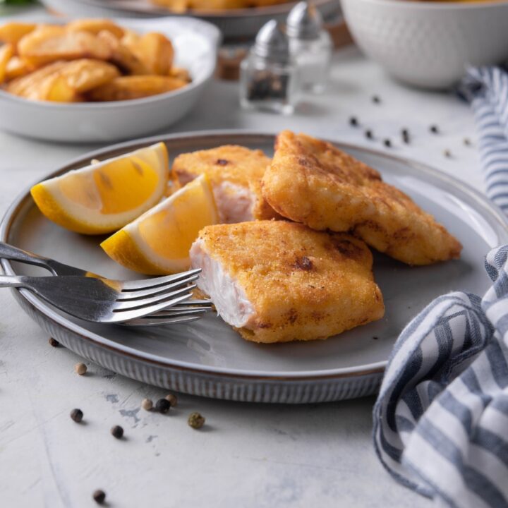 Fried grouper fillets sliced in half and piled on top of each other. The fish is on a blue plate alongside a set of silverware and two lemon wedges. In the background are plates of more fried fish and lemons.
