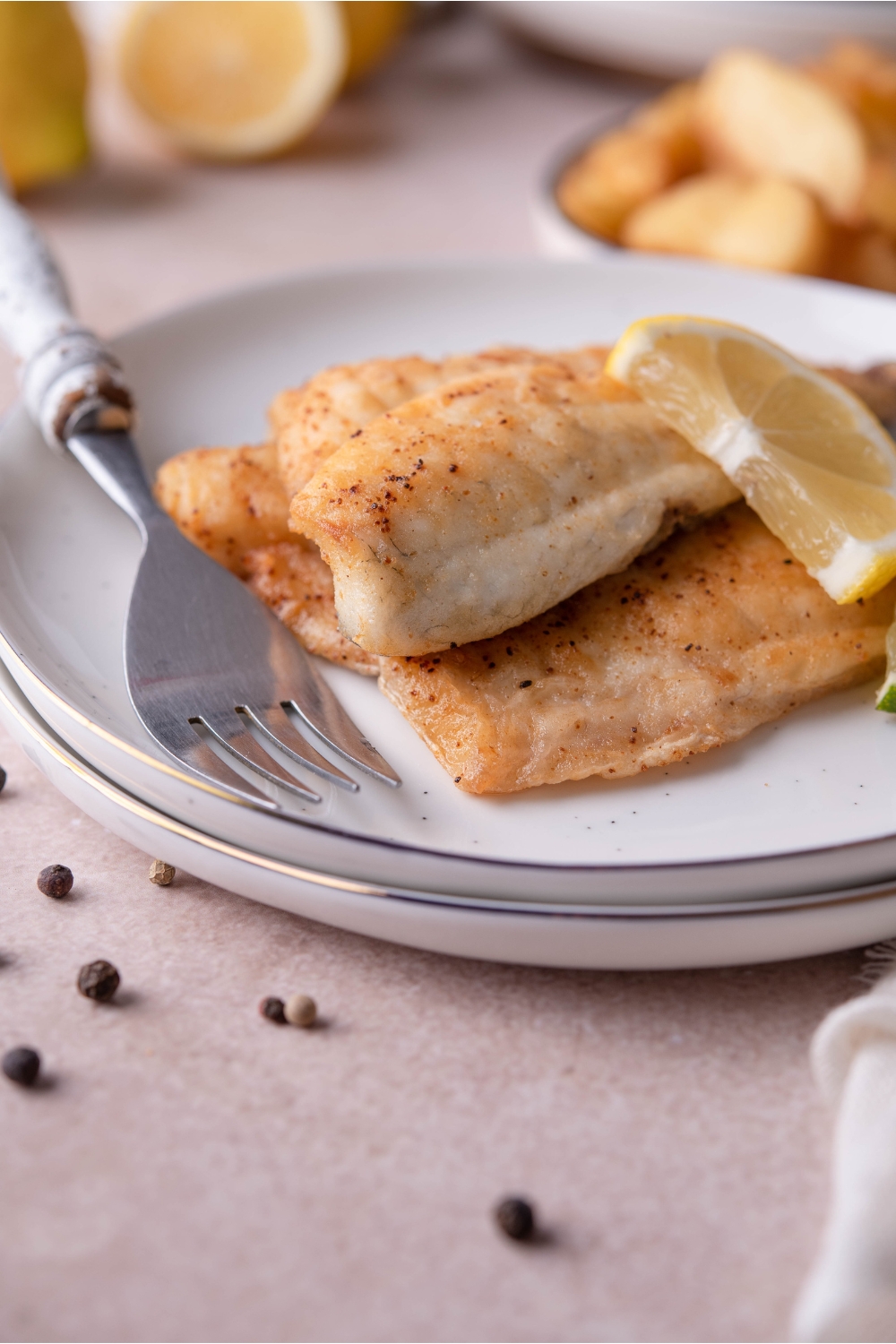 Two pan-fried tilapia fillets plated on top of each other with a slice of lemon on the fish. There is a fork on the plate and a second white plate under the plate of fish.