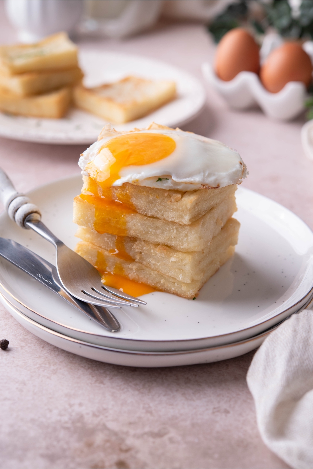 Four grit cakes stacked evenly on top of each other with a sunnyside up egg and the yolk oozing over the grit cakes. There is a set of silverware on the plate.