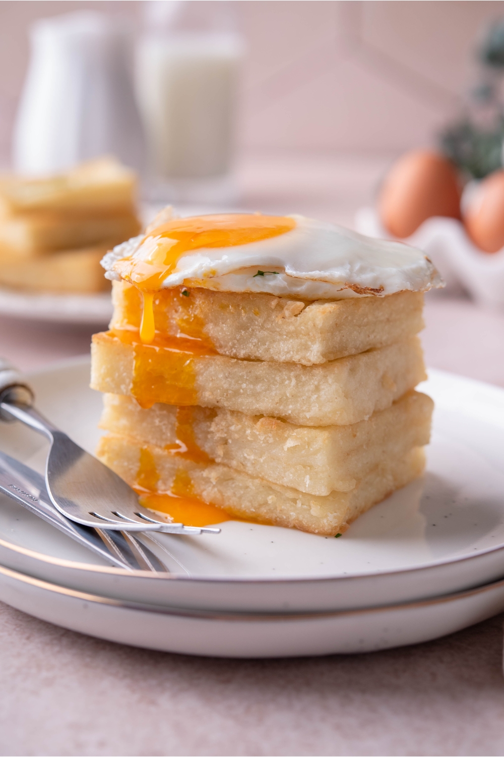 Four grit cakes stacked evenly on top of each other with a sunnyside up egg and the yolk oozing over the grit cakes. There is a set of silverware on the plate.