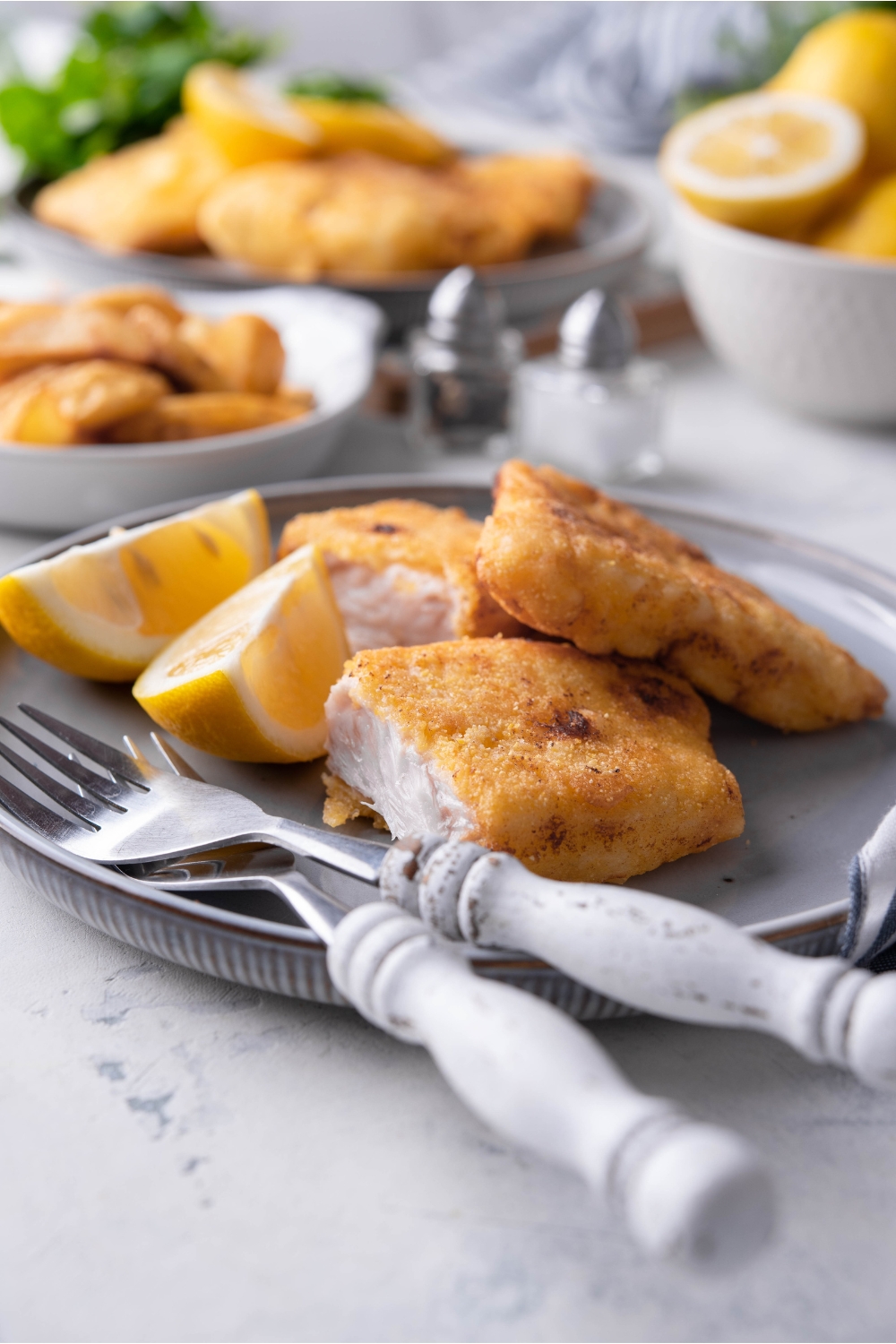 Fried grouper fillets sliced in half and piled on top of each other. The fish is on a blue plate alongside a set of silverware and two lemon wedges. In the background are plates of more fried fish and lemons.