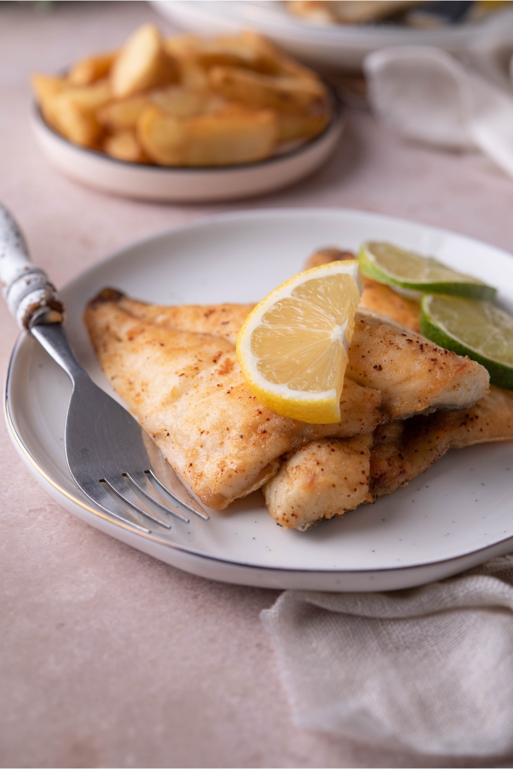 Two pan-fried tilapia fillets plated on top of each other on a white plate, each garnished with a sliced lemon and sliced lime. There is a fork on the plate and a plate of potato wedges in the background.