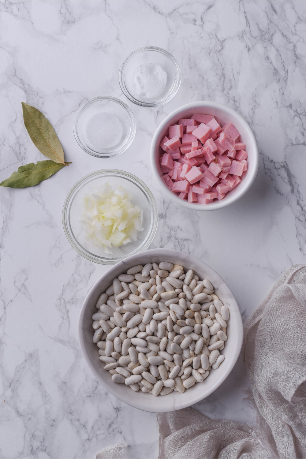 An assortment of ingredients including bowls of dried beans, cubed ham, diced onion, salt, and two bay leaves.