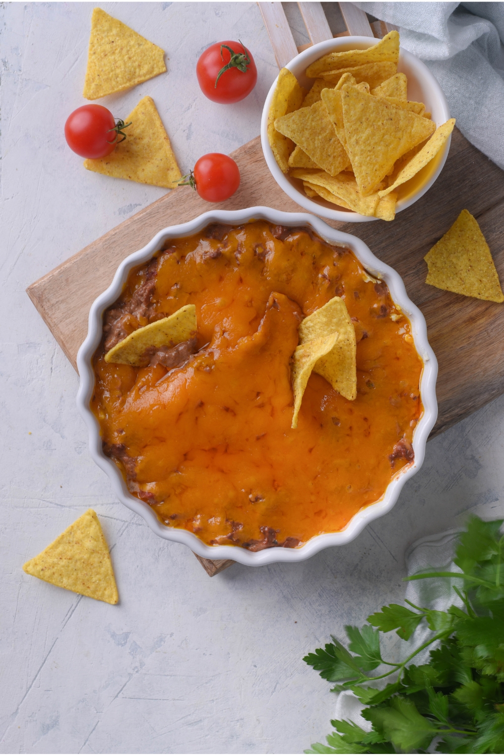 Overhead view of skyline chili dip with three tortilla chips sticking out of the dip. The dip is on a wood board and next to it are cherry tomatoes and a bowl of tortilla chips with some sprinkled on the counter.