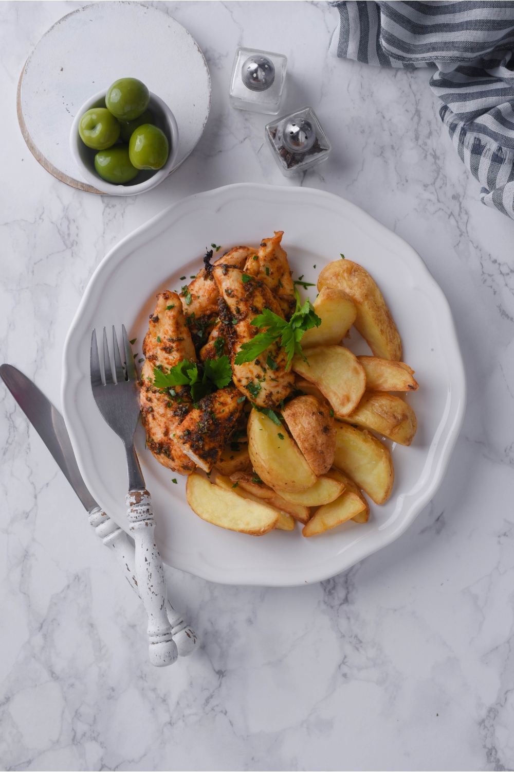 Baked chicken tenders on a white plate garnished with fresh herbs and served with a side of potato wedges. Surrounding the plate of chicken tenders are a fork, knife, salt and pepper shakers, and a small bowl of olives.