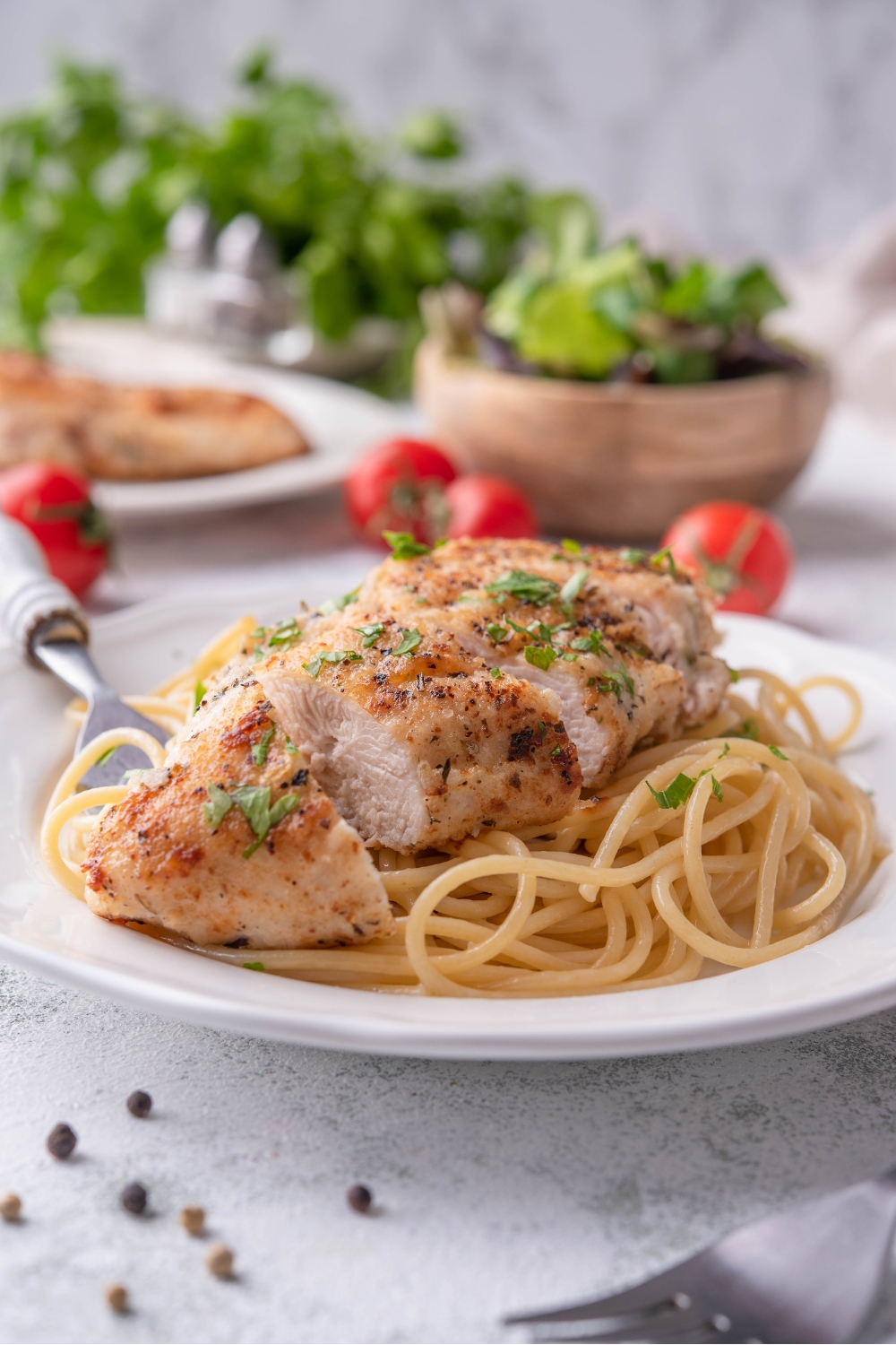 A sliced pan-fried chicken breast atop a bed of spaghetti noodles on a white plate with a fork on the plate. The chicken is garnished with fresh green herbs.