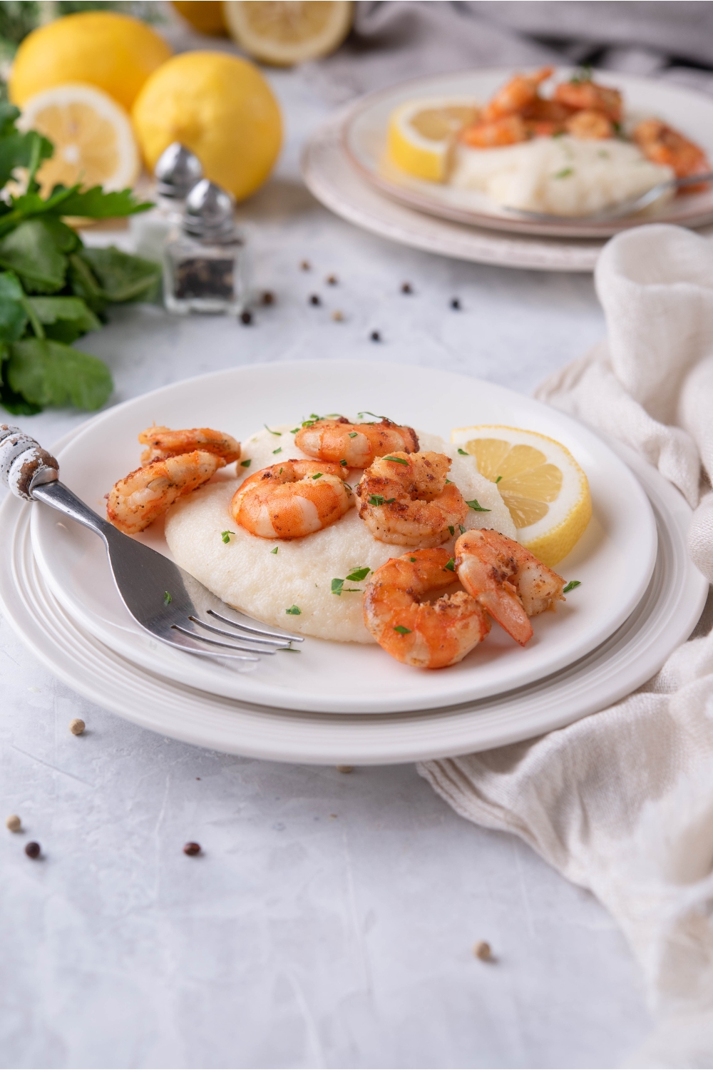 Pan seared shrimp atop grits with a garnish of fresh herbs and a lemon wedge. The plate of shrimp is on a second plate and there is a fork on the plate. In the background are more lemons and a second serving of shrimp.