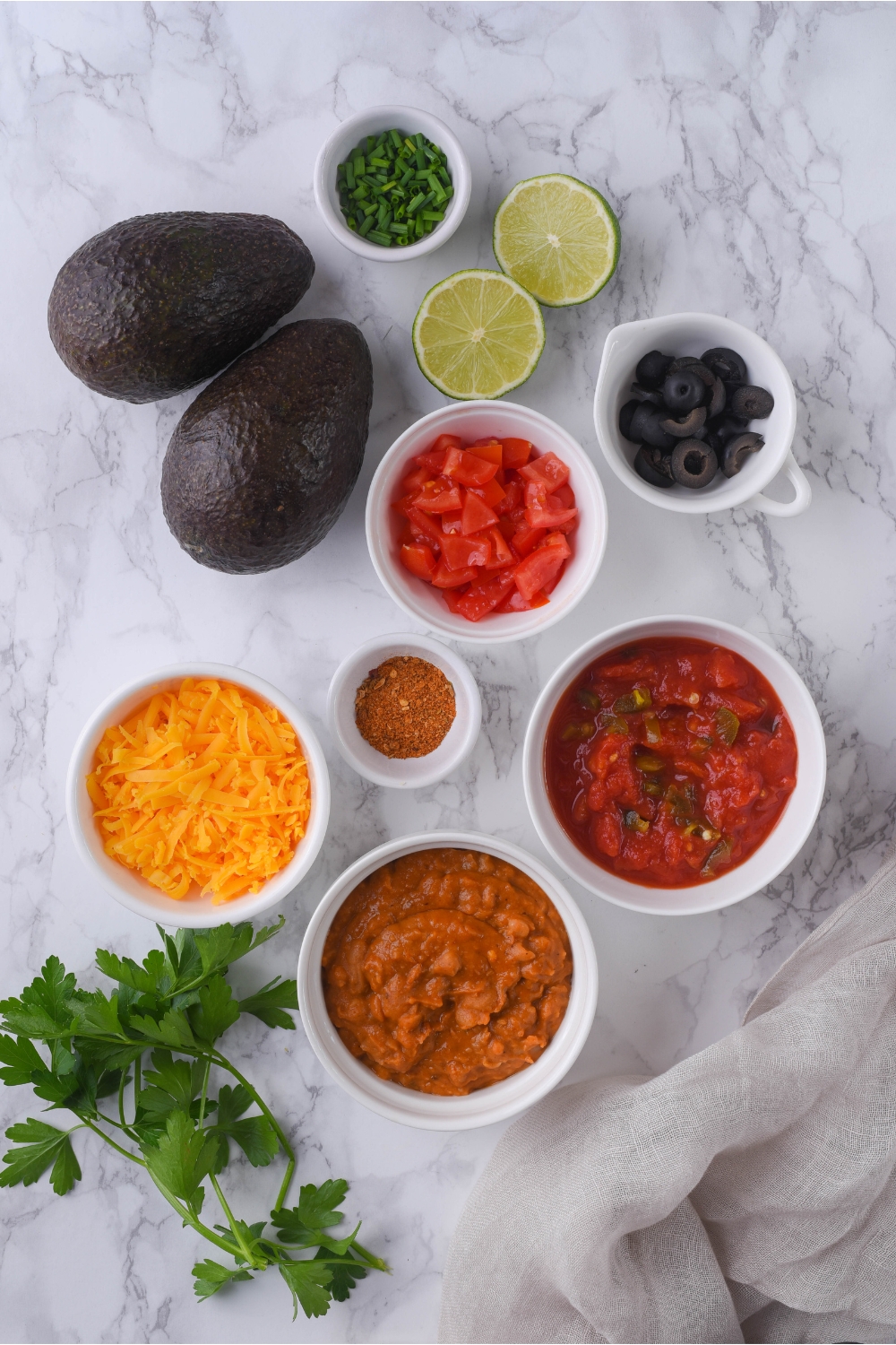 An assortment of ingredients for 7 layer dip including bowls of salsa, shredded cheese, beans, black olives, spices, diced tomatoes, and two avocados.