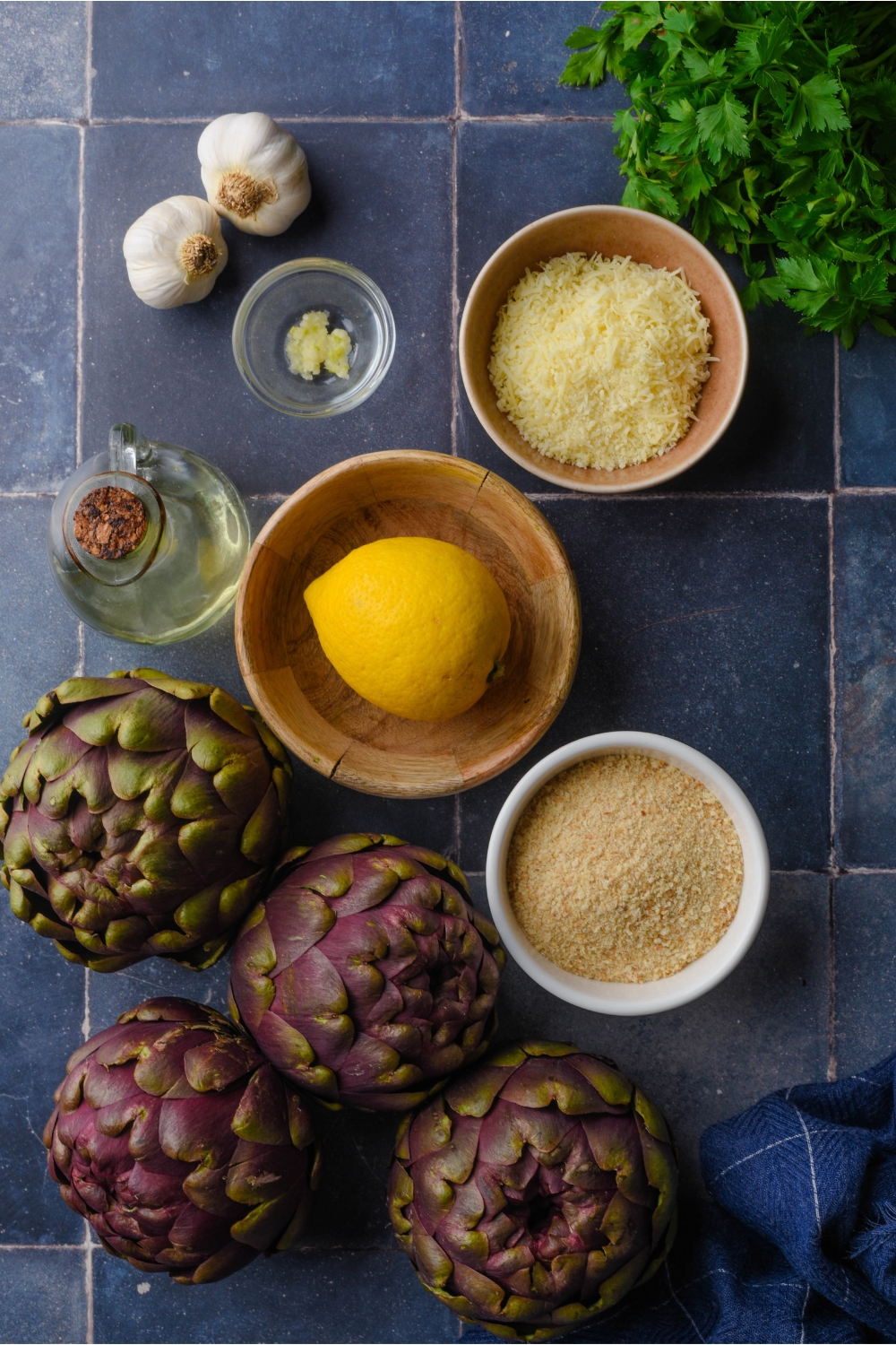 An assortment of ingredients including four whole artichokes and bowls of bread crumbs, parmesan cheese, garlic, one whole lemon, and a bottle of oil.