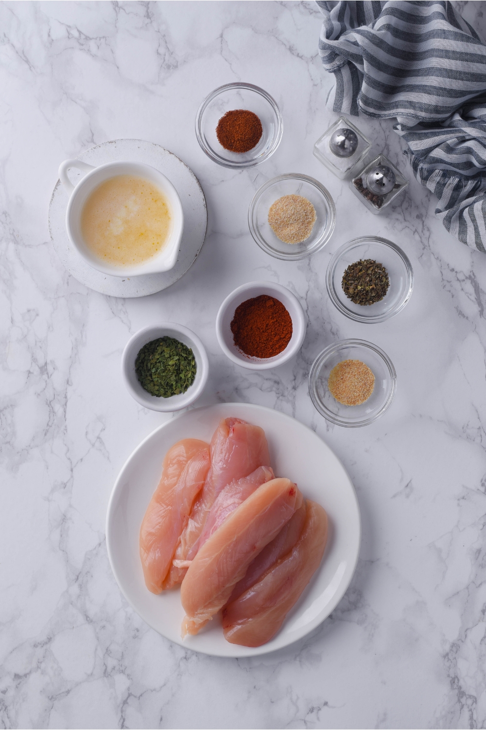 An assortment of ingredients for baked chicken tenders including raw chicken tenderloins, spices, and melted butter.