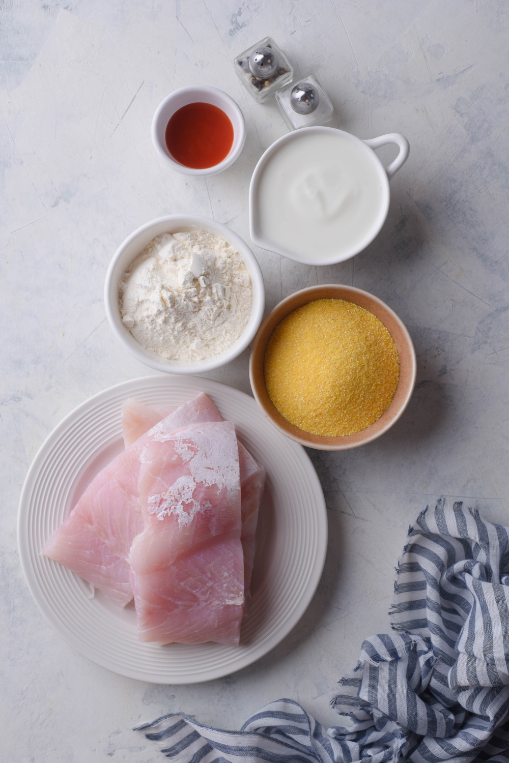 An assortment of ingredients including a plate of raw grouper and bowls of cornmeal, flour, buttermilk, and spices.