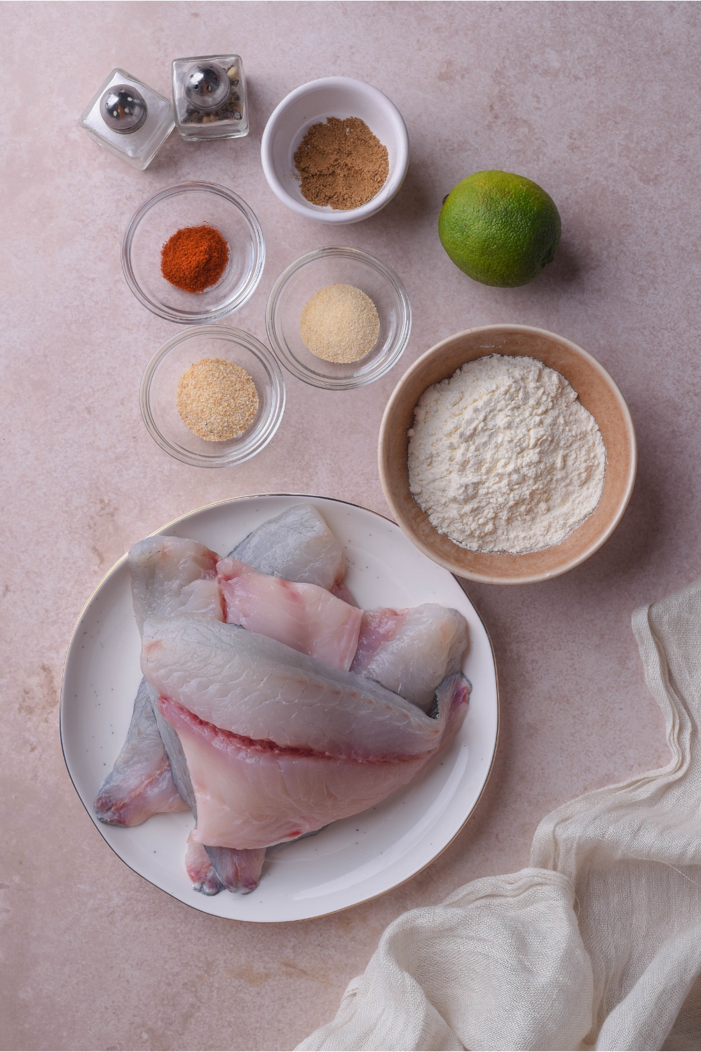 An assortment of ingredients including a plate of tilapia fillets and bowls of flour, spices, a lime, and salt and pepper shakers.