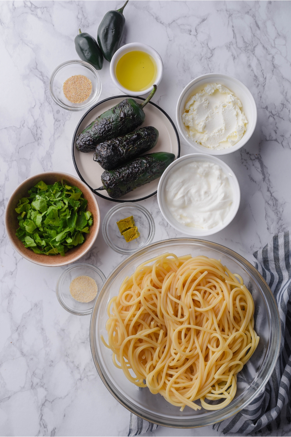 An assortment of ingredients including bowls of cream cheese, sour cream, diced green onion, cooked spaghetti noodles, and a plate of roasted jalapeños.