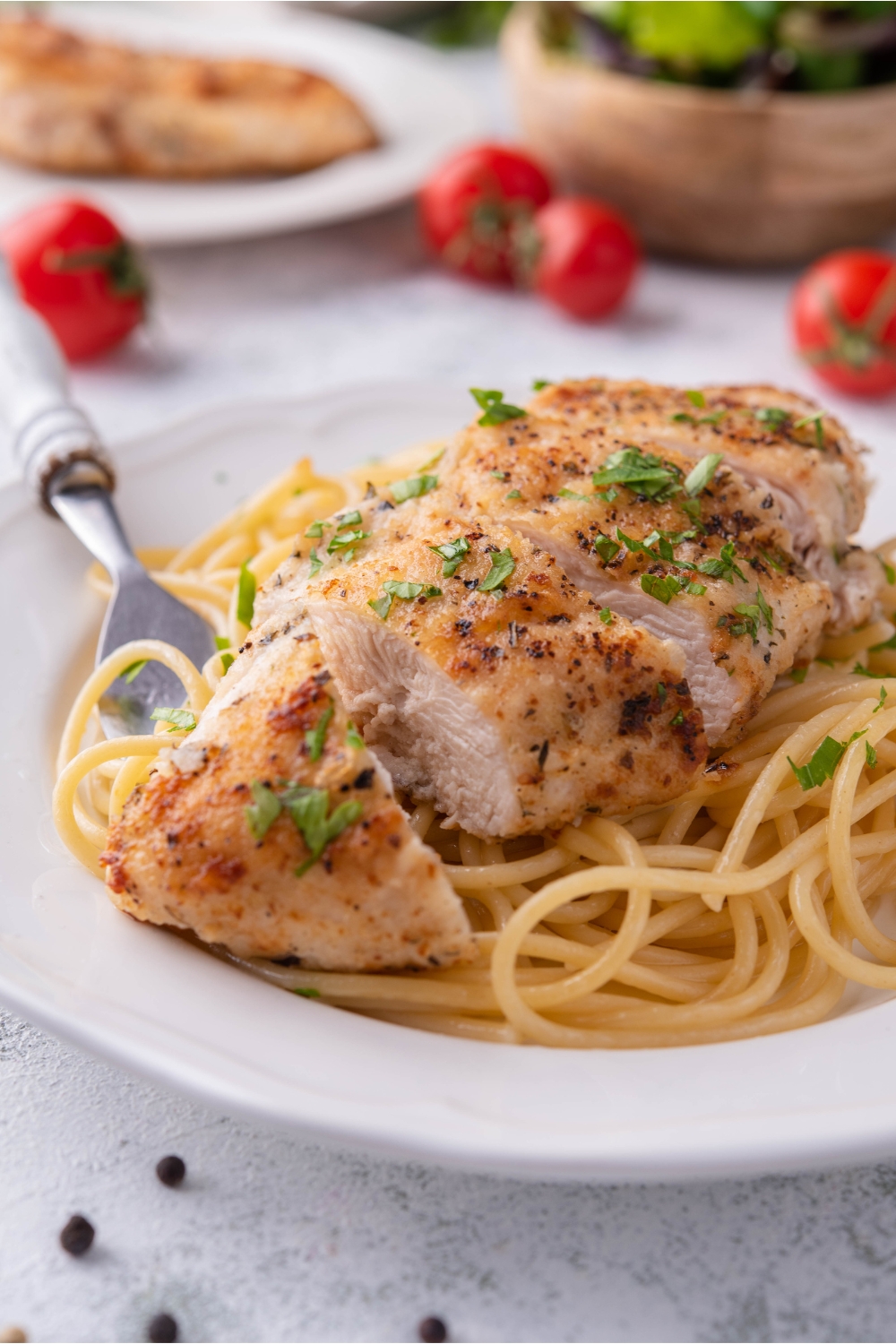 A sliced pan-fried chicken breast atop a bed of spaghetti noodles on a white plate with a fork on the plate. The chicken is garnished with fresh green herbs.