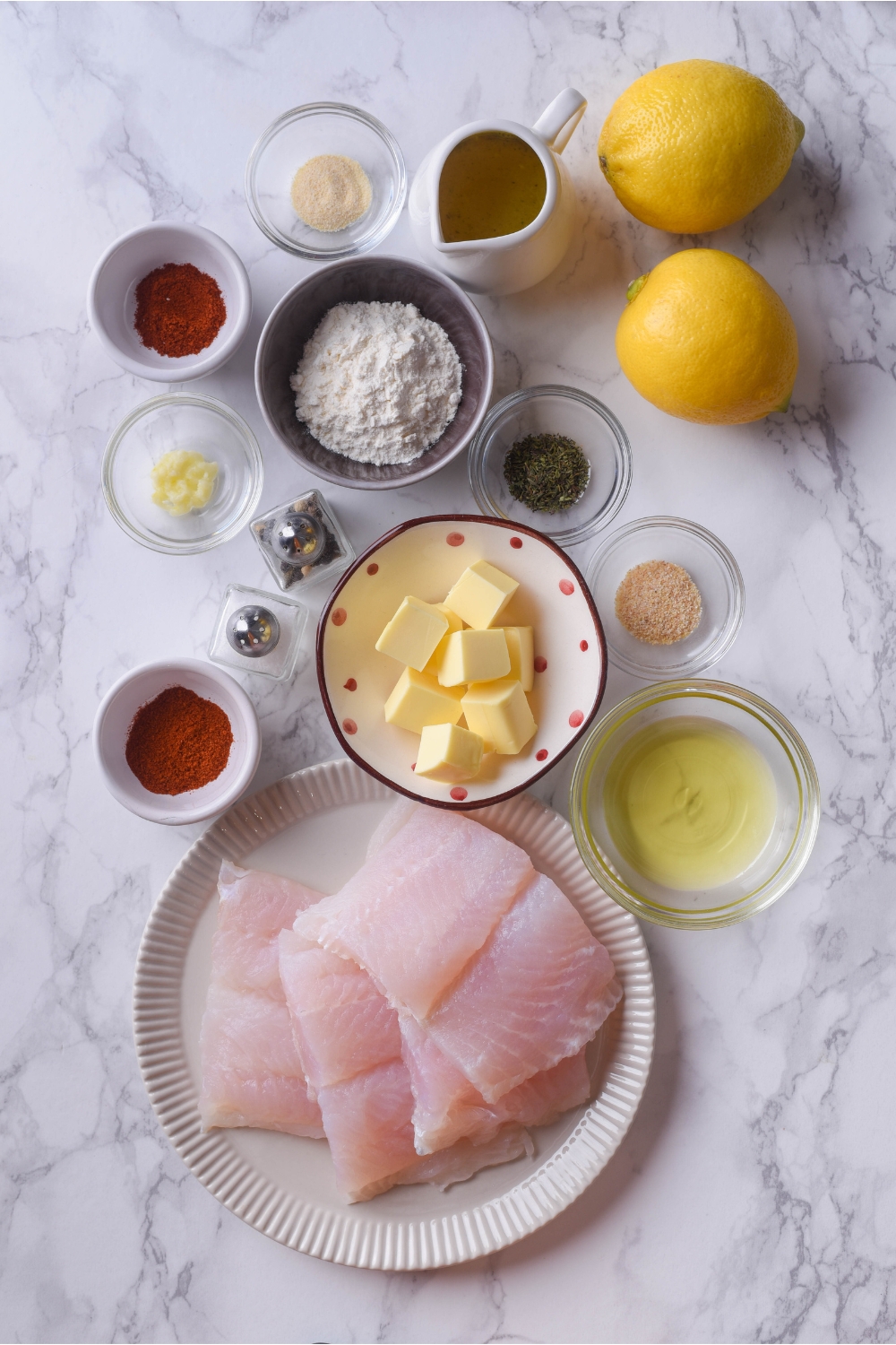 An assortment of ingredients including a plate of raw cod and bowls of butter, oil, spices, garlic, and two whole lemons.