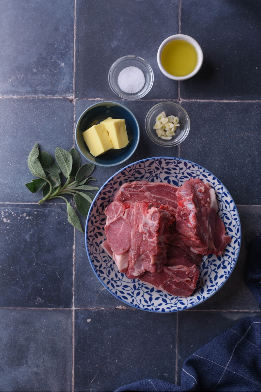 An assortment of ingredients including a plate of raw lamb and bowls of oil, salt, garlic, butter, and a sprig of fresh sage.