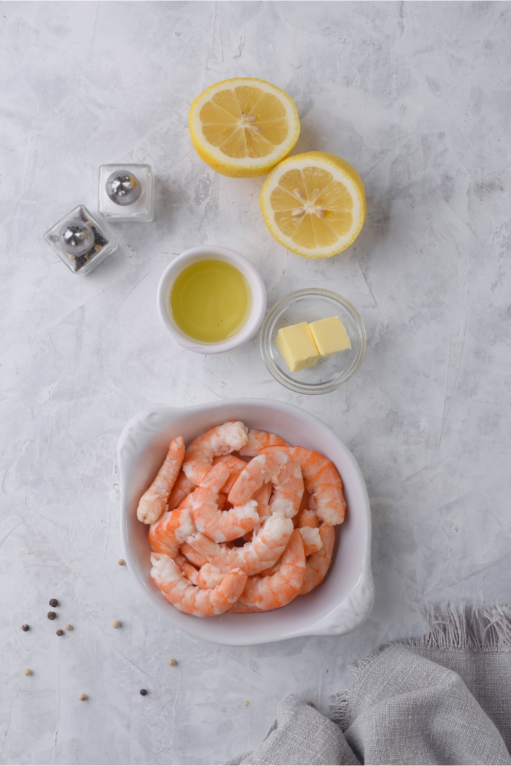An assortment of ingredients including a bowl of shrimp, smaller bowls of butter and oil, salt and pepper shakers, and a lemon cut in half.