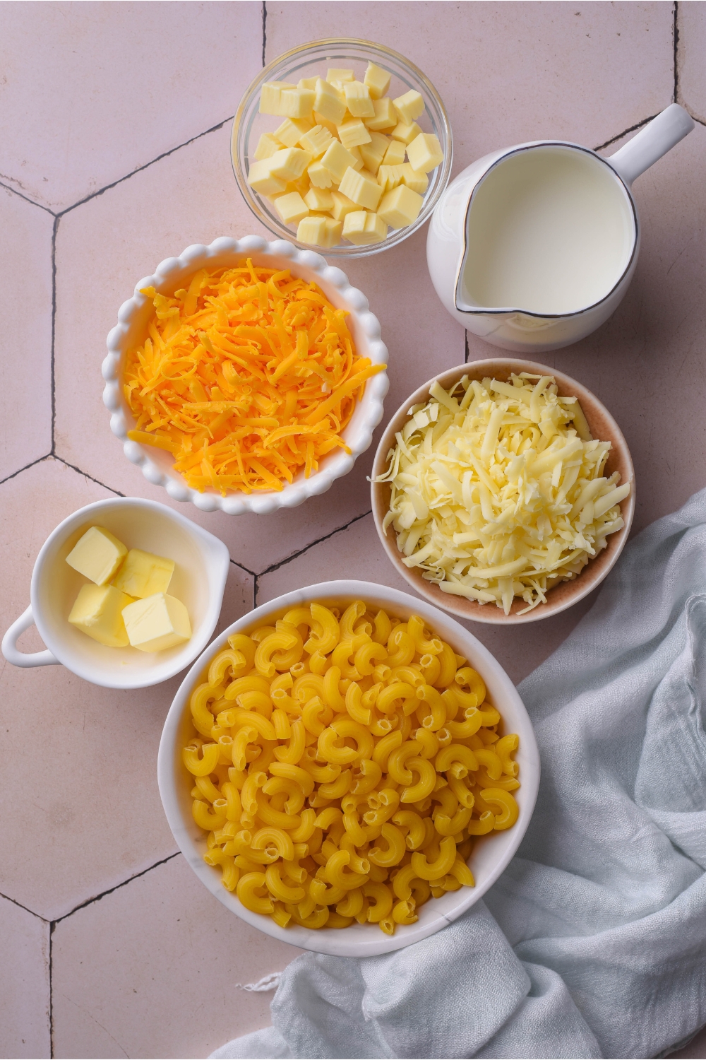 An assortment of ingredients including bowls of elbow macaroni, shredded cheese, butter cubes, and milk.