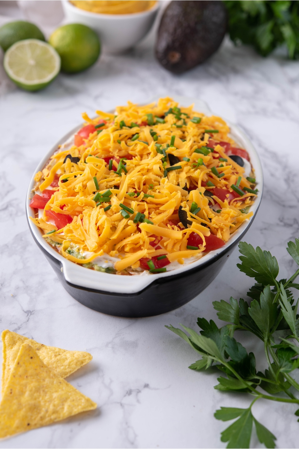 7 layer dip in a black and white baking dish, surrounded by tortilla chips and ingredients for making 7 layer dip.