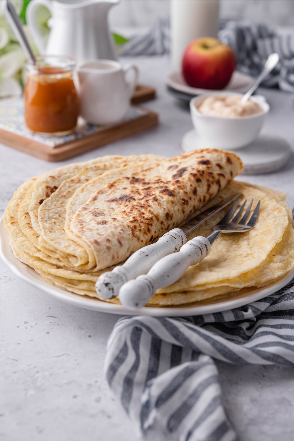 A stack of lefse flatbread with half of the stack folded on top of itself and a fork and knife on the stack of lefse. In the background is an assortment of fillings and toppings for the lefse.
