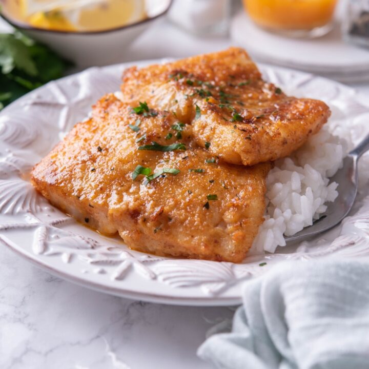 Two pan-seared cod fillets stacked atop a bed of white rice on a white plate with a fork on the plate. In the background is a second serving of cod and an assortment of ingredients.