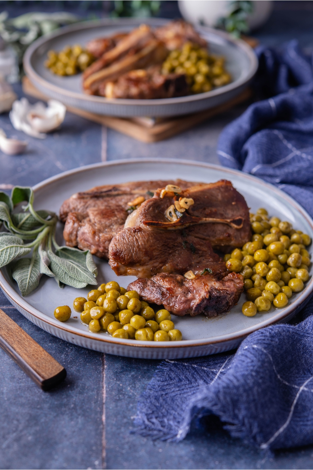 Pan seared lamb chops on a blue plate with a side of cooked peas. There's a garnish if fresh sage on the plate. Next to the plate is a set of silverware and there is a second plate of lamb chops in the background.