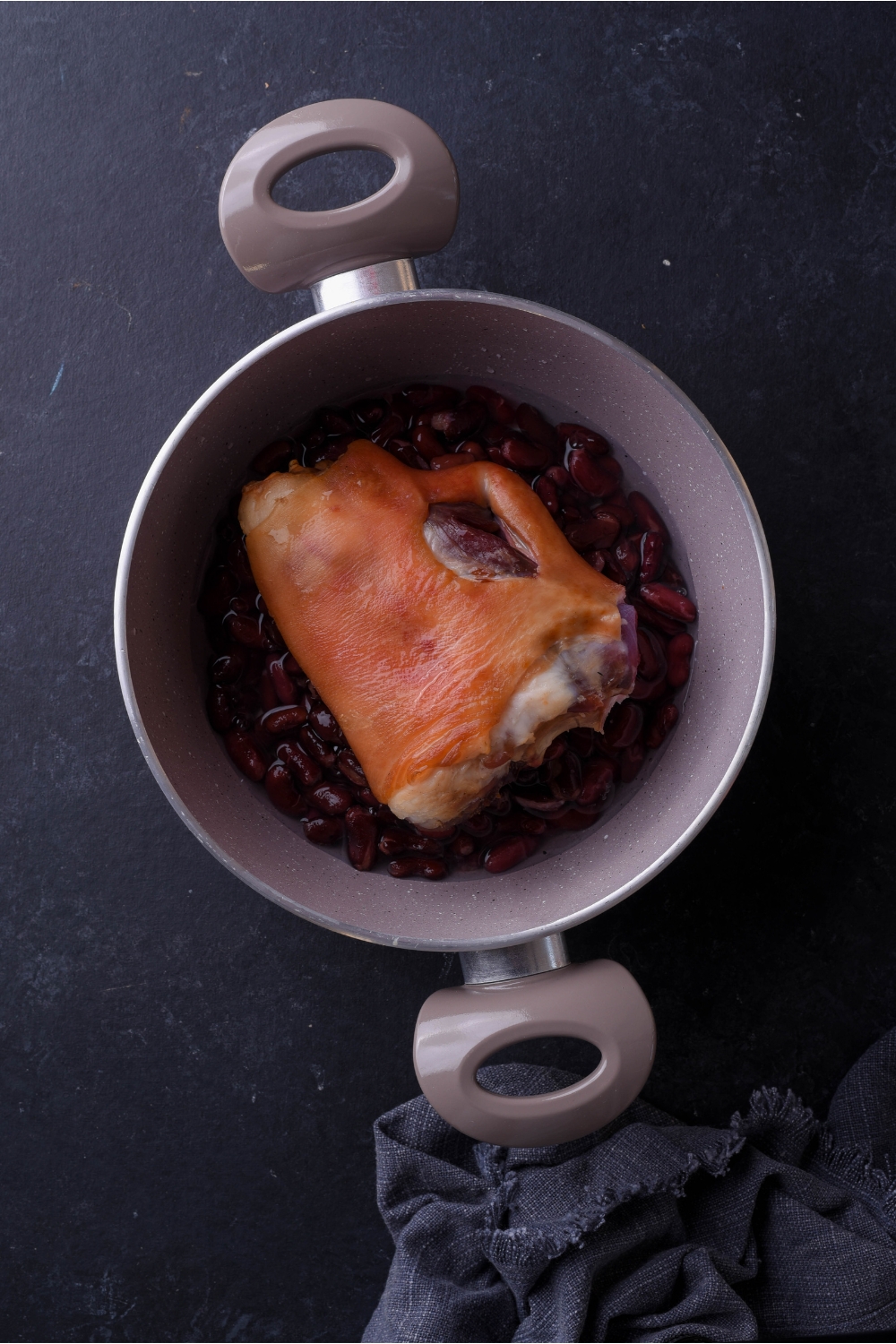 A grey saucepan filled with beans with a ham hock on top of the beans.