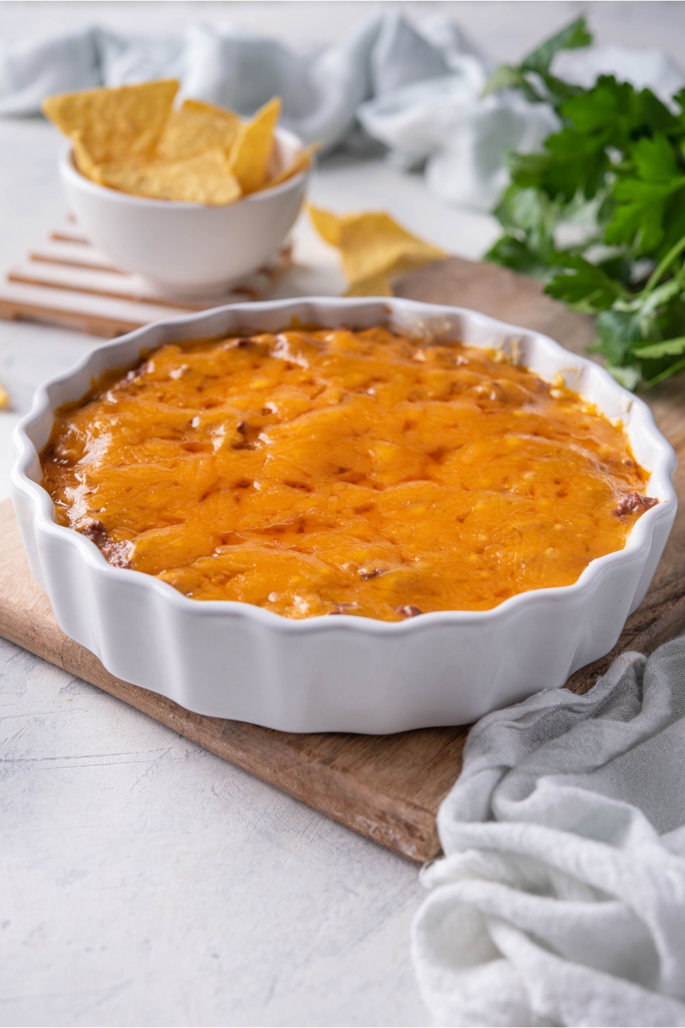 Skyline chili dip in a white serving dish covered in melted cheese. The chili dip is on a wood board and in the background is a bowl of tortilla chips and fresh herbs.