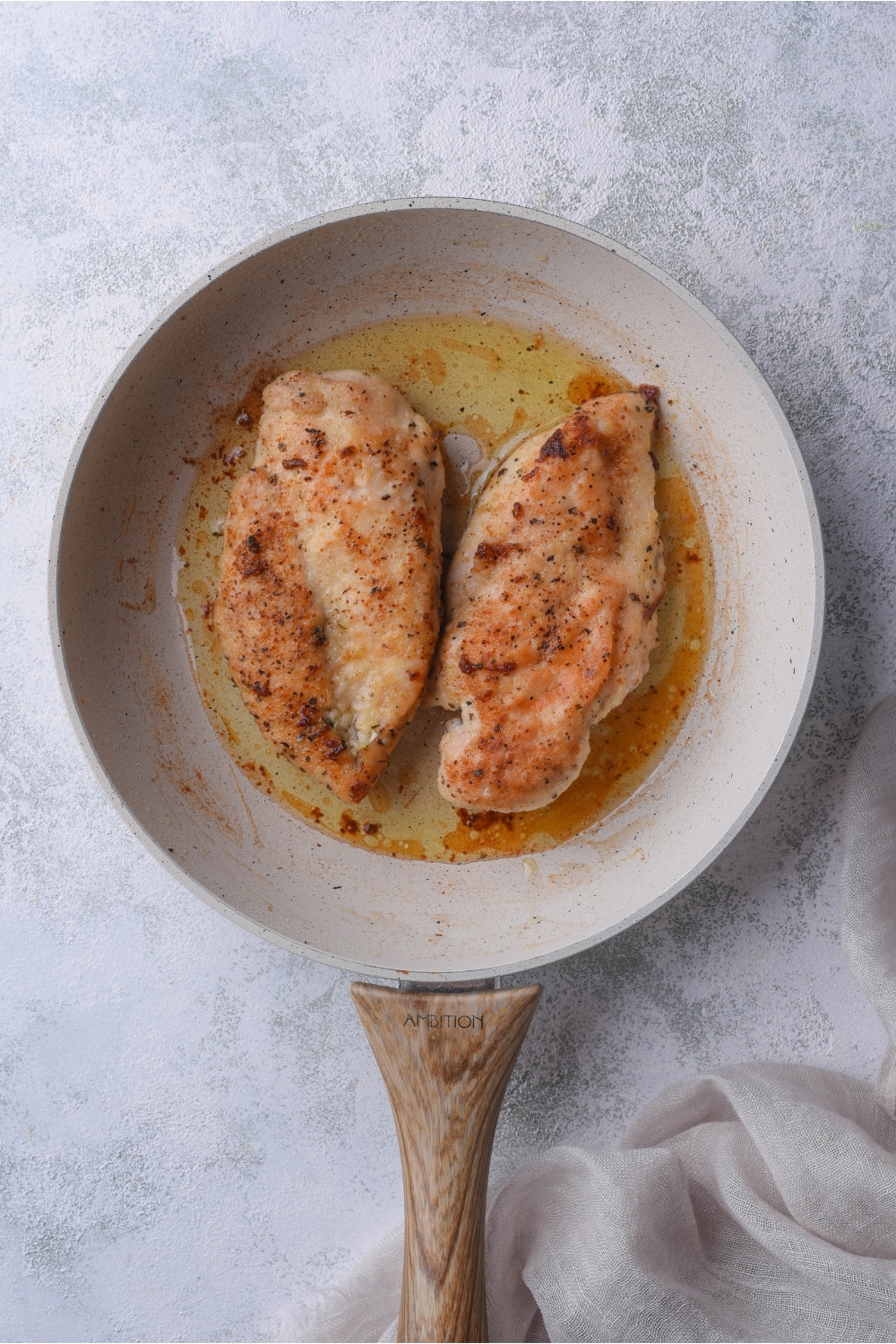 A grey skillet with a wooden handle and two seasoned and pan-fried chicken breasts in it.